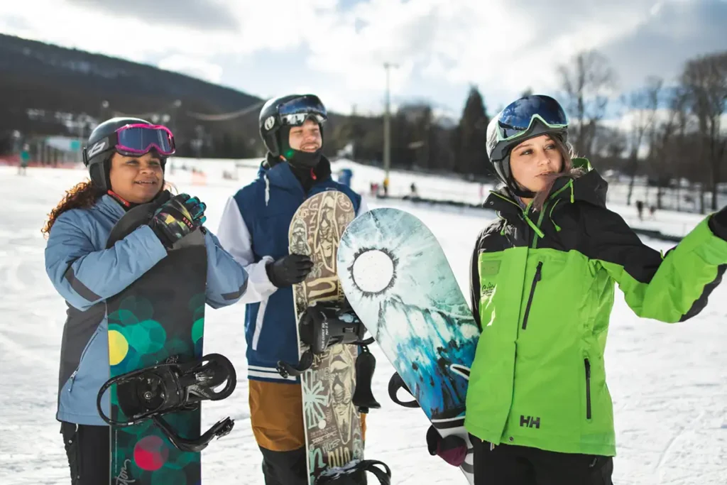 A group of three snowboarders dressed in winter gear stand on a snowy slope, holding their snowboards while chatting and looking at the surroundings. One woman in a bright green jacket gestures towards the distance, while the others smile and listen.